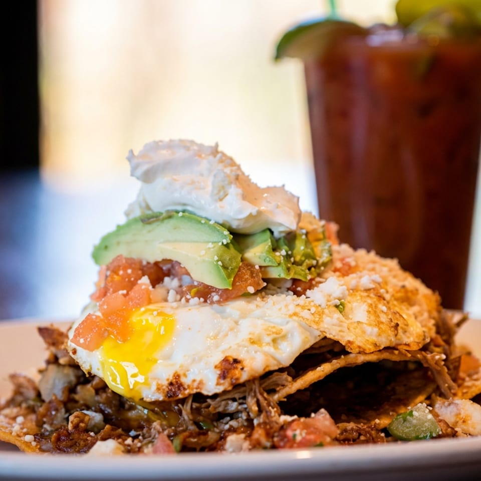 Stacked Mexican-style breakfast with fried eggs, avocado, crispy tortillas, salsa, and a garnished tomato drink.
