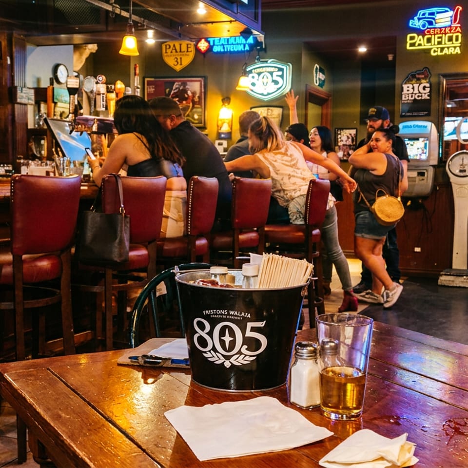 Lively pub interior with patrons at the bar, beer bucket, drinks, neon signs, and red leather stools around a wooden table.