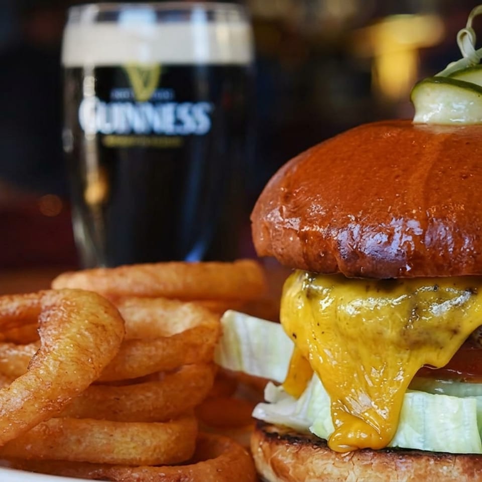 Cheeseburger with lettuce, tomato, pickles, and melted cheddar beside a stack of onion rings on a plate.