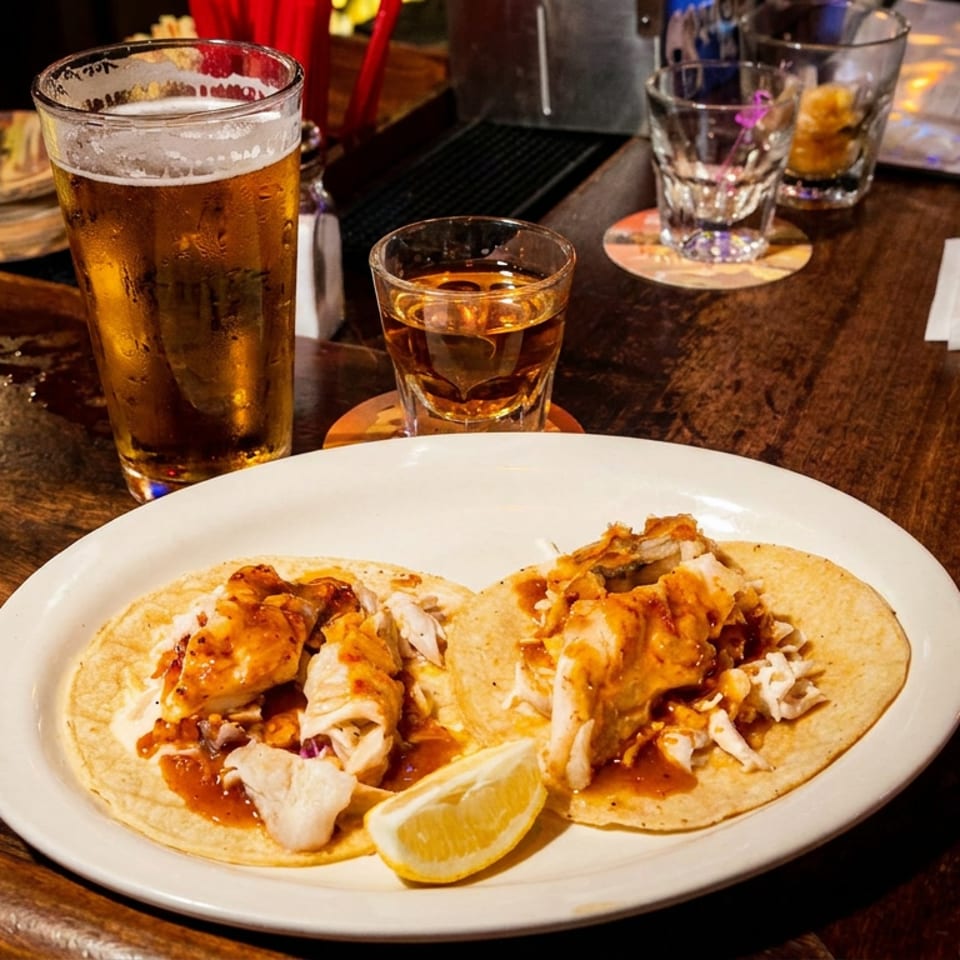 Two saucy fish tacos with shredded cabbage and lemon wedge sit on a plate at a dimly lit bar beside beer and whiskey.