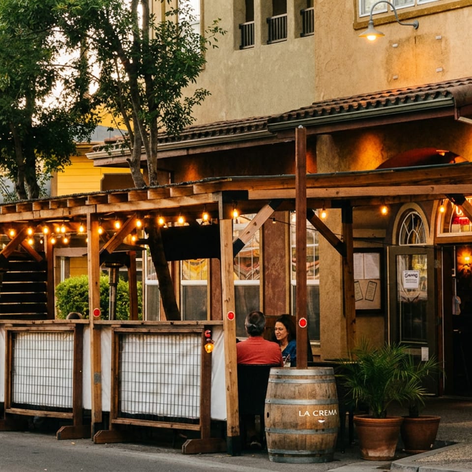 Rustic restaurant patio with string lights, wooden covered seating, potted flowers, and two diners outside a stucco building.