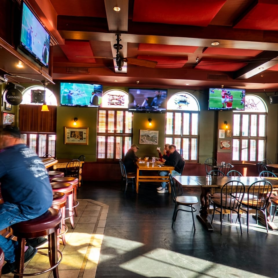 Warmly lit sports bar with a long wooden counter, patrons, dining tables, televisions, and colorful beer signs.