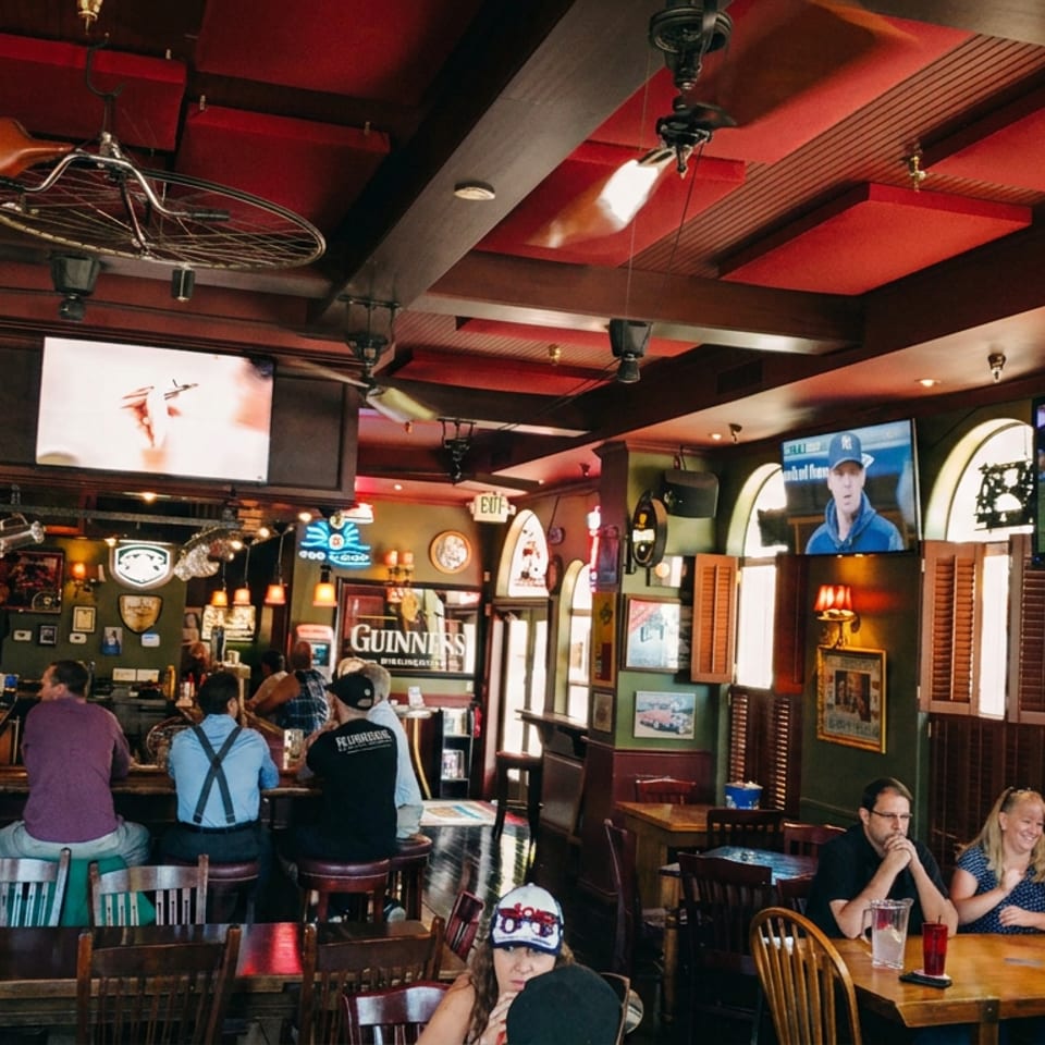 Bustling Irish-style pub interior with patrons at the bar and tables, Guinness signage, sports televisions, and dark wood decor.